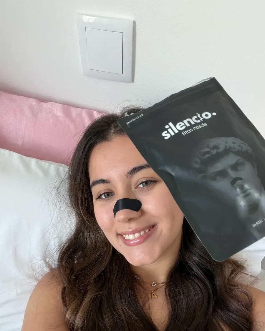 Criadora deitada na cama com uma fita nasal posta, a segurar a embalagem do produto ao lado da cara.
